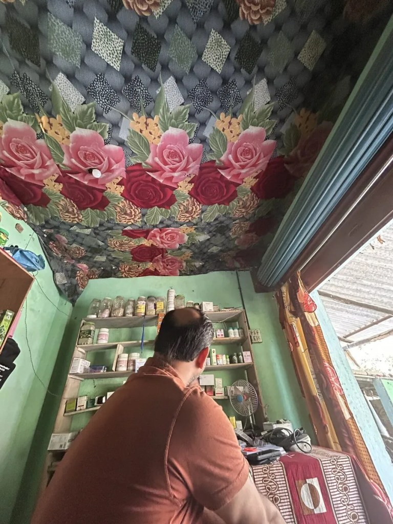 Seated in a village medicine shop in Purulia, seen from behind, facing shelves of medicines